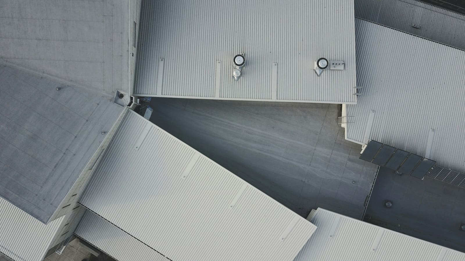 Overhead drone view of industrial building roofs, with ribbed metal panel runs beside darker low-slope sections and roof vents.