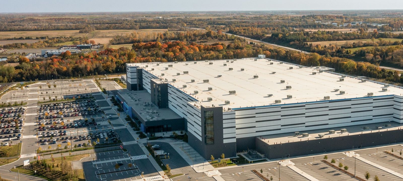 Aerial view of a large distribution building with a wide white low-slope roof field, rooftop units spaced across it and a parking lot alongside.