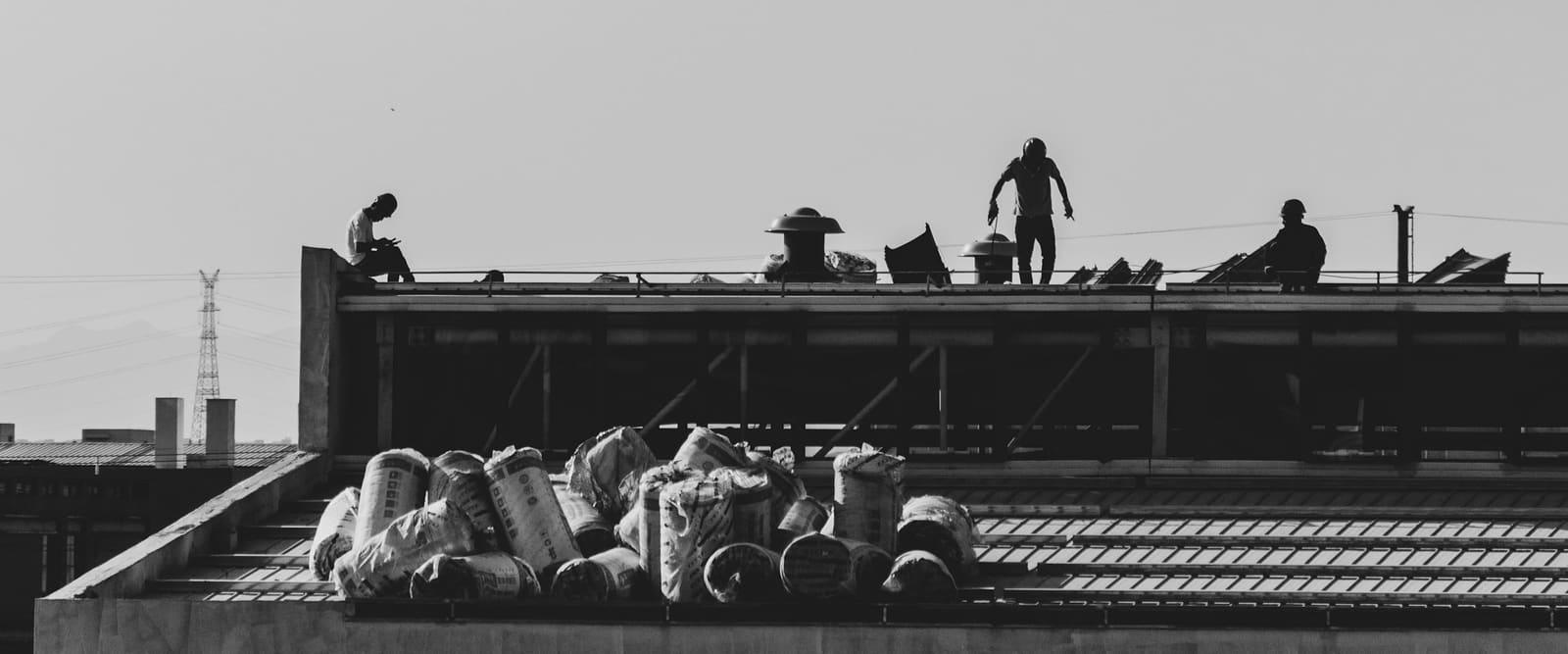 Roofing crew working along the edge of a flat commercial roof, with mushroom vents in the field and bundles of material staged on the deck below.